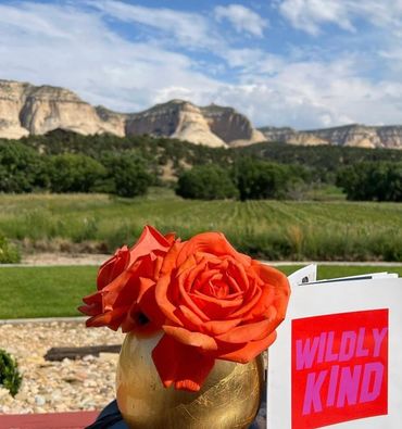 Wildly kind tour, Bright orange roses in a golden vase with scenic mountains and greenery in the background.