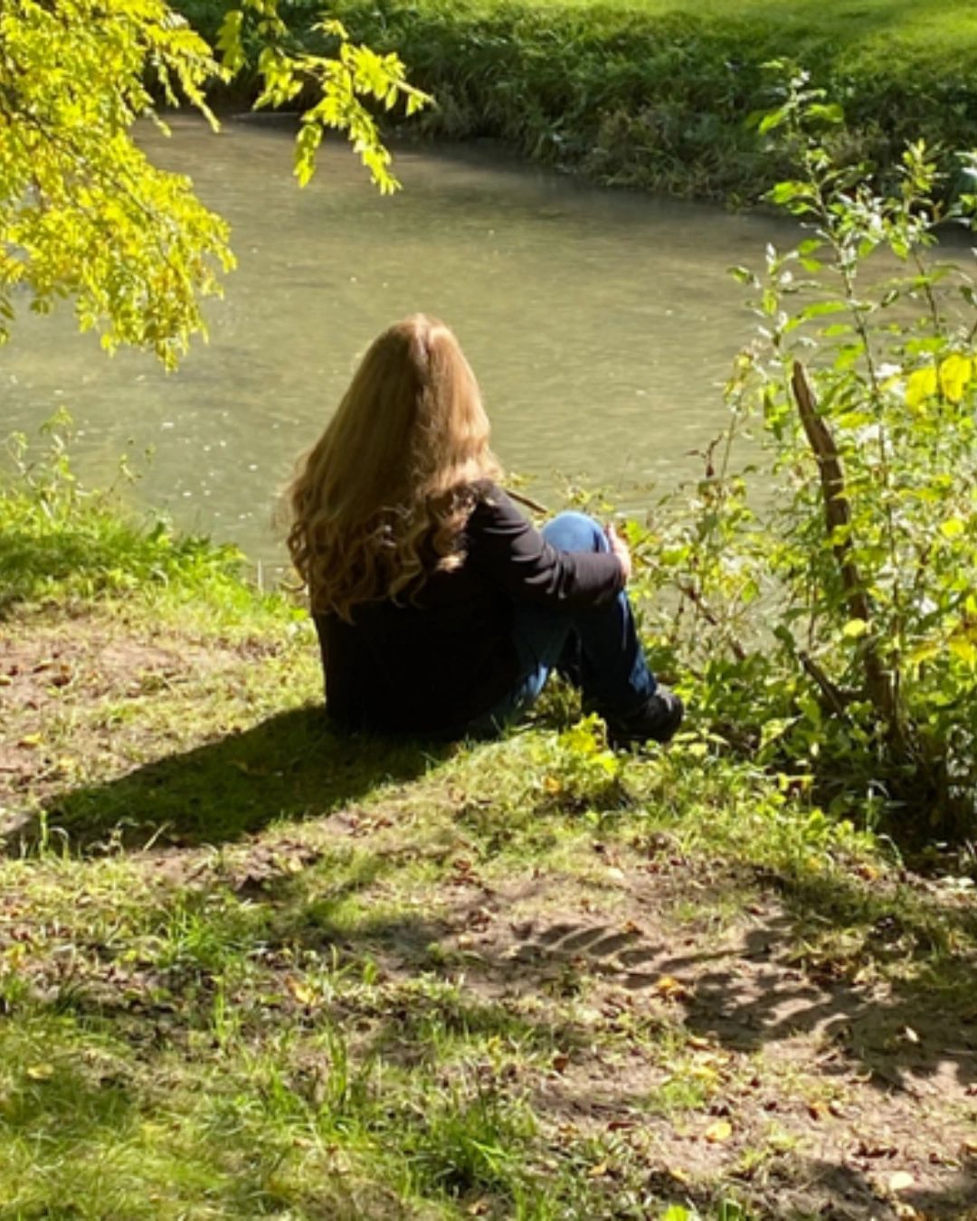 Woman sitting by a calm river surrounded by greenery in a peaceful outdoor setting