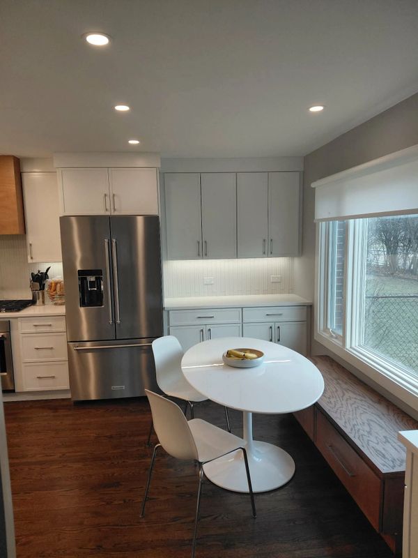 Modern kitchen with white cabinets, stainless steel fridge, and a small dining table by a large window.
