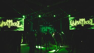 Concert scene with green lighting and crowd in front of stage screens.