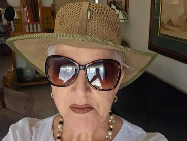 Elderly woman wearing a large sun hat and oversized sunglasses indoors.
