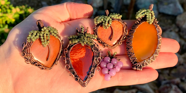 Hand holding wire-wrapped pendants that are shaped around stones that represent different fruits