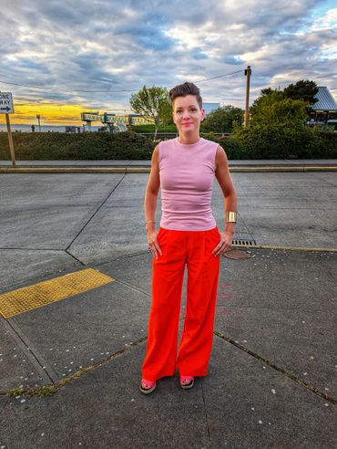 A woman in a pink top and red pants stands confidently on a city sidewalk at sunset.