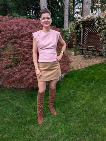 Woman standing on grass wearing a pink top, beige skirt, and brown boots.