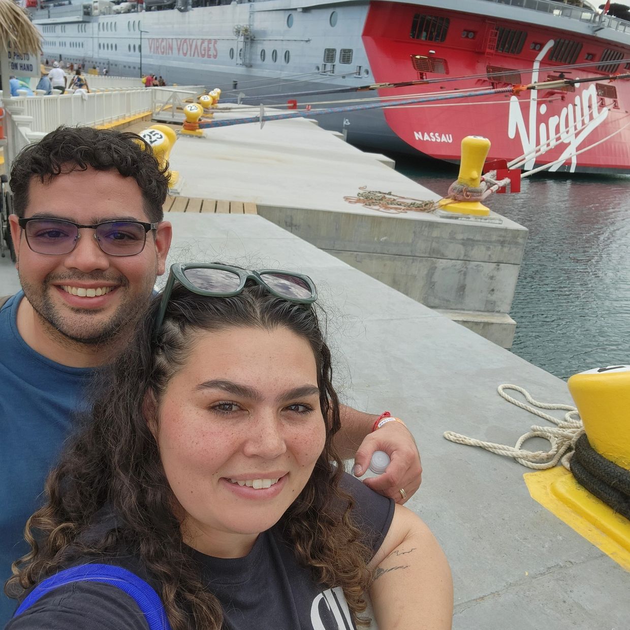 Smiling couple takes a selfie in front of a Virgin Voyages cruise ship docked at the pier.