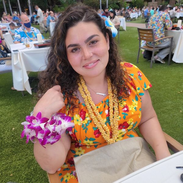 Woman in a floral dress with leis and a flower in her hair at an outdoor dinner.