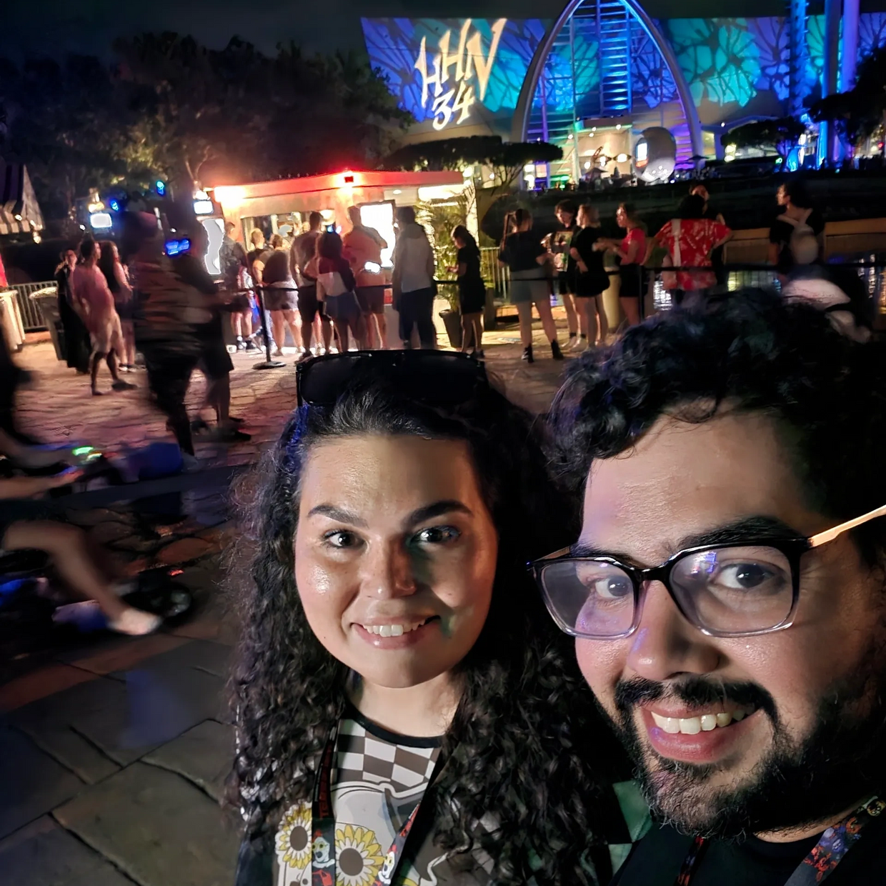 Couple smiling at night in front of a lit-up event with people in line.