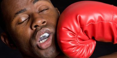 A boxer, wearing a mouthguard, braces as a red-gloved punch lands squarely on his mouth