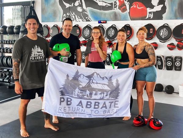 Group of five people holding a PB Abbate Return To Base flag in a boxing gym.