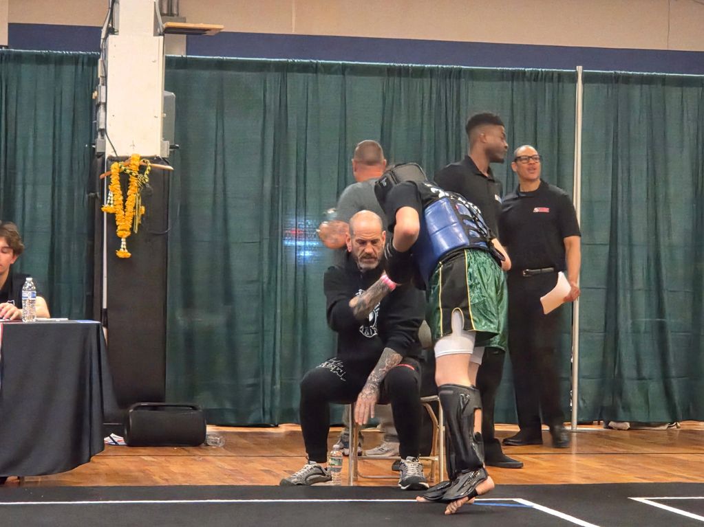 A martial artist in protective gear receives guidance from a seated coach.