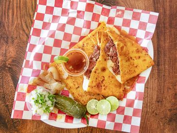 A quesadilla with lime slices, pickled vegetables, and sauce on checkered paper.