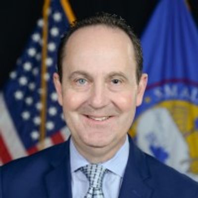 Smiling man in a suit with American and organizational flags in the background.