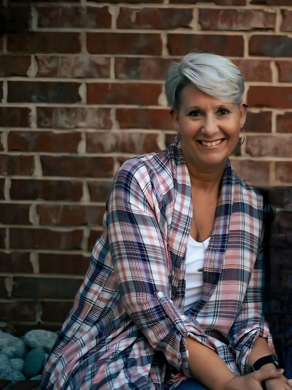Smiling woman with short gray hair wearing a plaid shirt against a brick wall.