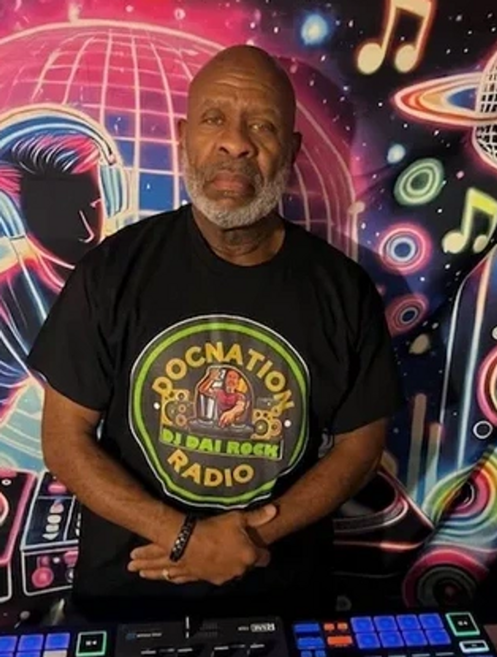 An older man with a beard wearing a DJ-themed t-shirt stands in front of a colorful music mural.