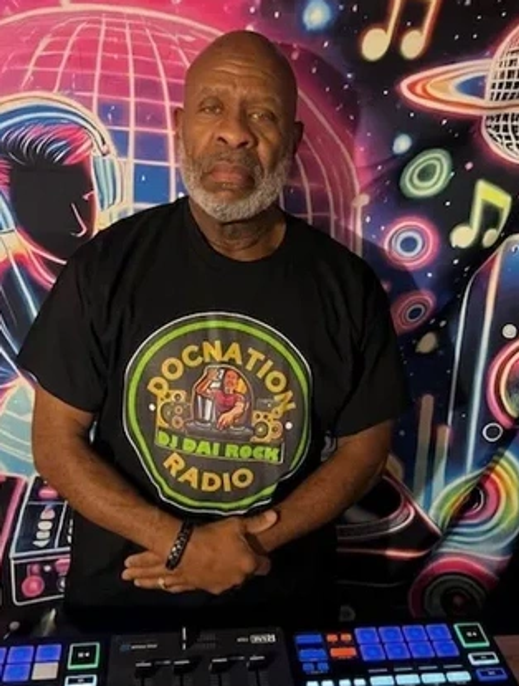 An older man with a beard wearing a DJ-themed t-shirt stands in front of a colorful music mural.