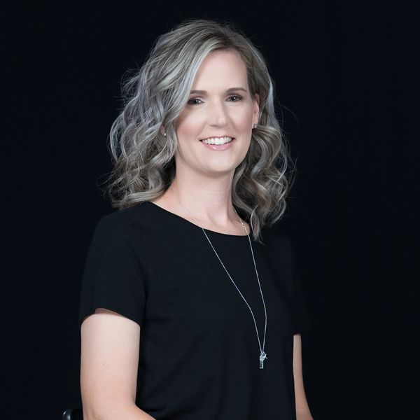 Smiling woman with curly gray hair wearing a black top and necklace against a black background.