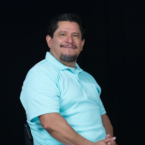 Smiling man in a light blue polo shirt sitting against a black background.