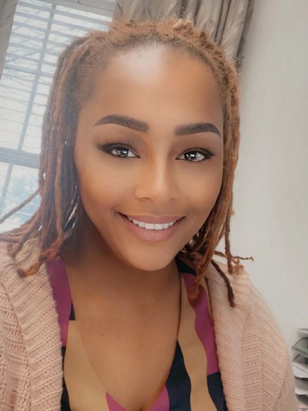 Smiling woman with dreadlocks wearing a pink sweater and colorful top.