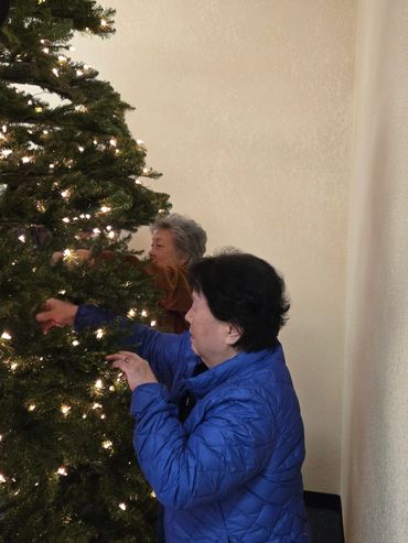 Two elderly women decorating a lit Christmas tree.