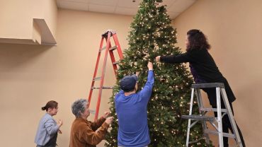 Four people decorate a large Christmas tree indoors.