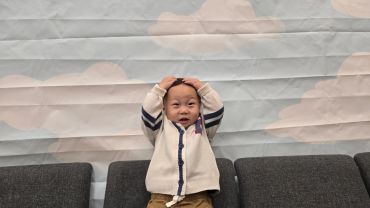 A joyful toddler with hands on head sitting on a couch against a cloud-patterned wall.