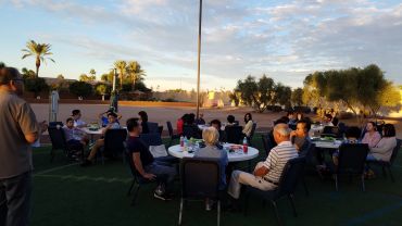 People gathered outdoors at sunset for a social event with round tables and chairs.