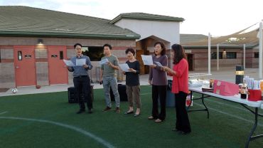 Five people standing outdoors, holding papers and singing together.