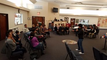 A person speaks to an attentive group seated in a classroom setting.