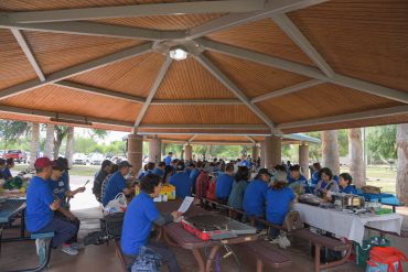 Group of people in blue shirts gathered under pavilion for a meal and meeting.