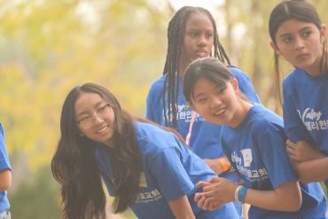 Group of young women in blue shirts outdoors, smiling and leaning forward.