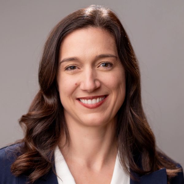Professional woman with brown hair smiling, wearing a navy blazer and white blouse.