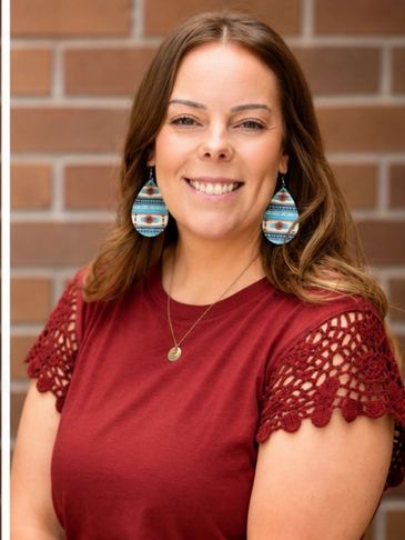 Smiling woman in a red top with patterned earrings against a brick wall.
