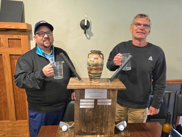 Two men holding mugs standing beside a trophy with golf tees.