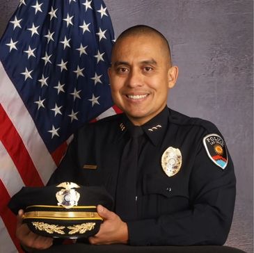 A smiling police officer in uniform posing with his hat in front of an American flag.