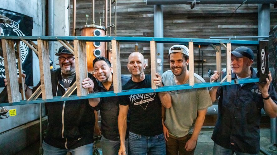 Five men smiling and posing behind a ladder in an industrial setting.