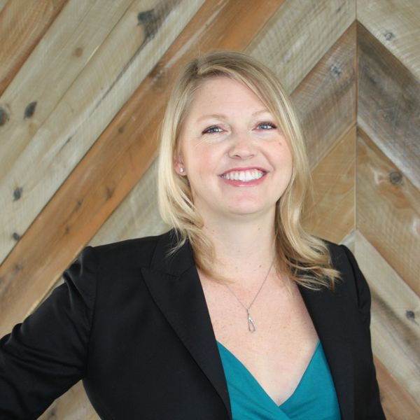 Smiling woman with blonde hair wearing a black blazer and teal top against a wooden background.