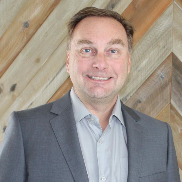 A smiling man in a gray suit and light blue shirt in front of a wooden wall.