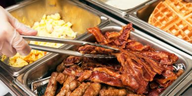 Buffet setup with bacon, sausages, scrambled eggs, and waffles.