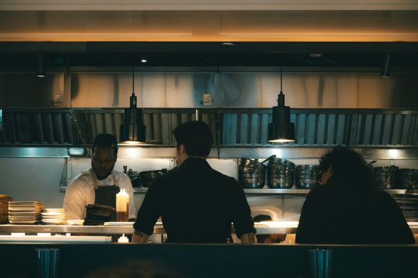 Three people in a restaurant kitchen, two facing the chef.