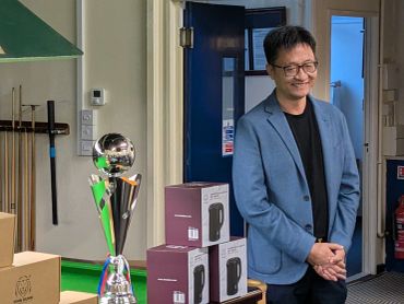 Man wearing glasses and blue blazer standing by a trophy and boxes.