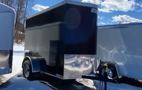 A black enclosed utility trailer parked on snowy ground under a partly cloudy sky.