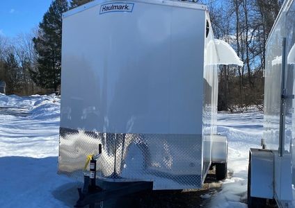 A white Haulmark trailer parked on snowy ground with trees in the background.
