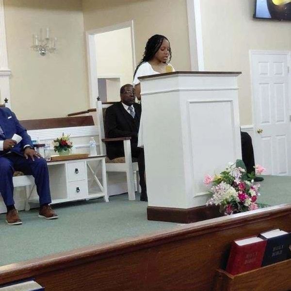 A young woman speaking at a church podium with seated men behind her.