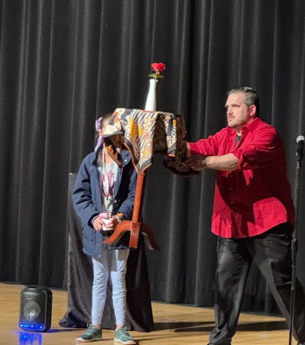 A man in red performs a magic levitation trick with a table and flower on stage.