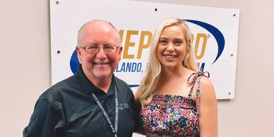 Two people smiling in front of a company sign.