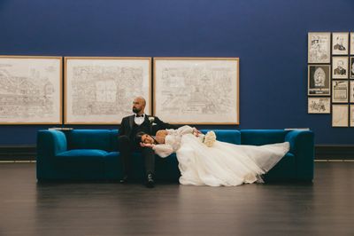 Bride in white gown lies on blue sofa beside groom in black suit in gallery.