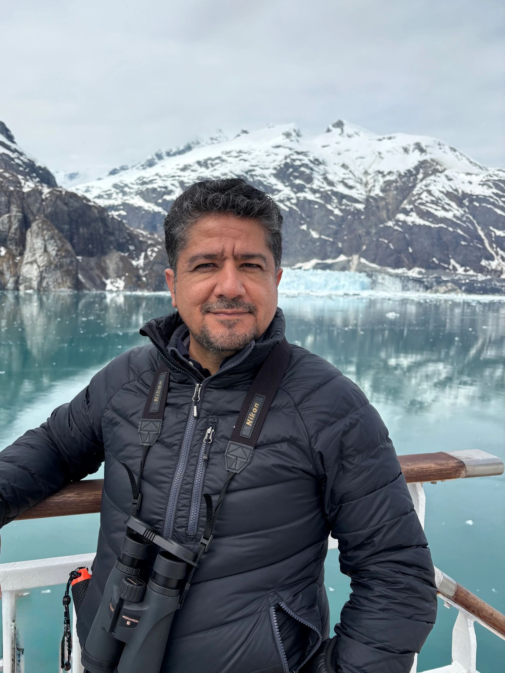 Man in a black jacket with binoculars, snowy mountains and icy lake in the background.