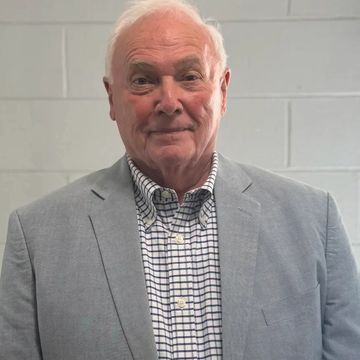 Elderly man in a light gray suit jacket and checkered shirt standing against a white brick wall.