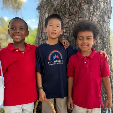 Three diverse boys smiling and standing together outdoors near a tree.