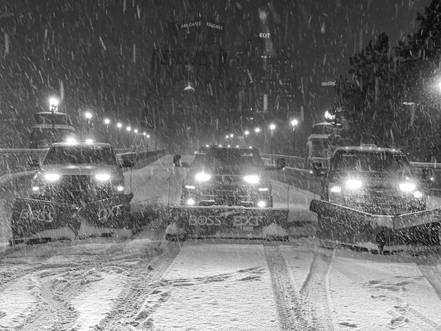Three snowplows clearing a snowy road at night with headlights on.